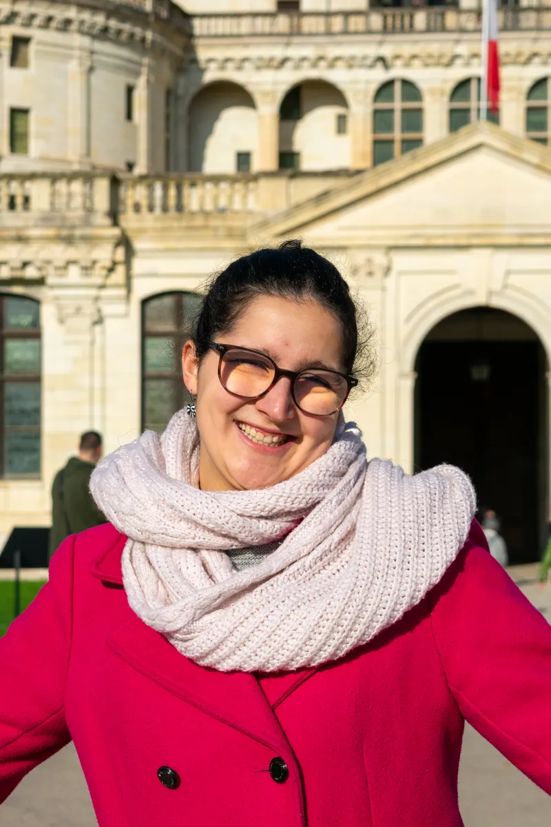 Moi, la guide, avec des lunettes, un manteau rose et une écharpe blanche devant un bâtiment de pierre blanche.